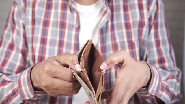 A male person in a plaid shirt and white t-shirt, holds an empty wallet with his hands, expressing financial problems, financial crisis, or poverty.