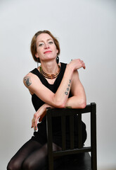Fototapeta premium Selbstbewusstes Frauenportrait im Studio. Model in schwarzem Outfit mit Tattoos, sitzend auf einem Stuhl – minimalistisches Setting mit klarer Haltung und moderner Ästhetik.