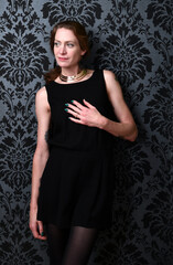 Naklejka premium Elegantes Frauenportrait im schwarzen Kleid vor ornamentaler Tapete. Selbstbewusste Pose mit dezenter Handbewegung – stilvolle Fashion-Inszenierung mit klassischer, dunkler Ästhetik.