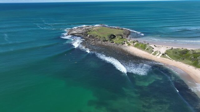 A small group of surfers wait in the lee of a beautiful headland, boards resting on gentle swells. Summer sun glows over a brilliant blue sea, capturred in smooth, sweeping drone footage.