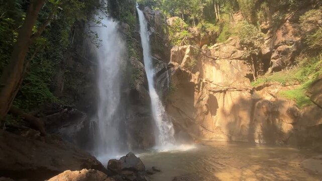 Twin waterfalls cascading down steep rocky cliff into misty natural pool surrounded by tropical forest and lush greenery creating dramatic landscape near Mok Fa Waterfall Thailand