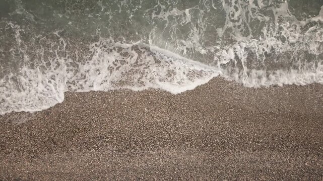 Loop-ready 4K UHD static overhead shot of foamy waves washing over a pebble beach shoreline; soothing natural texture for commercials, intros, wellness backgrounds.