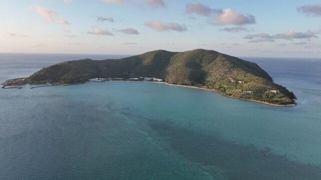 Slow drone pullback with a subtle rise at golden hour, widening over Hayman Island to reveal the turquoise bay, resort shoreline and sunlit green ridges, Whitsundays, Queensland, Australia.