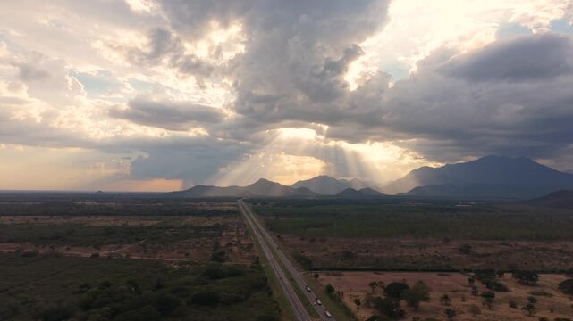 High-altitude drone shot pulling back over the straight Ruta del Sol highway, revealing the vast tropical landscape and dramatic crepuscular rays over the mountains of Cesar, Colombia, at dusk.