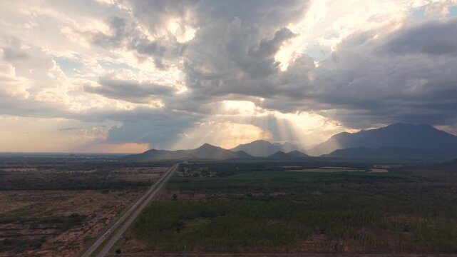 High-altitude drone shot pulling back-left over the straight Ruta del Sol highway, revealing the vast tropical landscape and dramatic crepuscular rays over the mountains of Cesar, Colombia, at dusk.