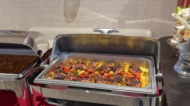 Indoor buffet catering setup with stainless chafing dish holding grilled chicken pieces over potatoes and sliced peppers, serving line food display under warm restaurant lighting.