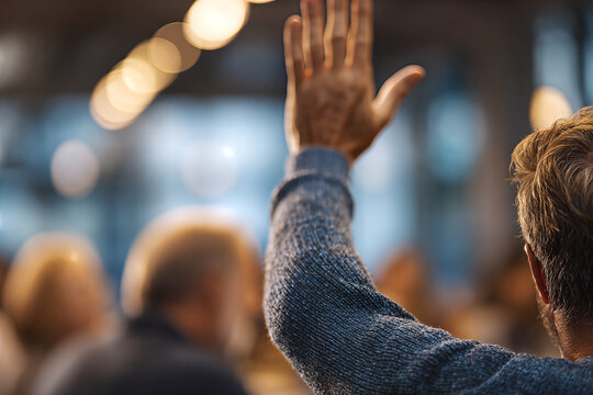Over - the - shoulder view of a middle - aged man in a blue knit sweater raising his hand to speak at a blurred corporate meeting with warm bokeh lights