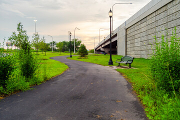 A quiet path curves beneath the overpass, where young trees, soft grass, and slender streetlamps create a gentle rhythm against the concrete. The scene feels like a small city&rsquo;s heartbeat