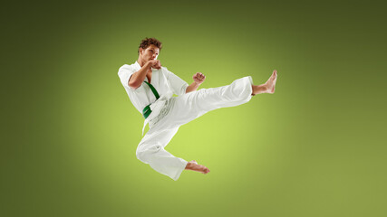 Fototapeta premium Young karate athlete delivering powerful side kick in mid air wearing white gi and green belt. Concept of explosive martial arts action, sport performance training and agility development.