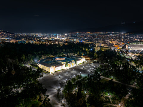 Aerial view of the illuminated building and parking lot surrounded by trees, with the city lights glowing in the distance, Athens, Greece.