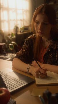 Vertical shot of red haired young woman using laptop while studying at home and twirling pencil thinking over assignments, scene lit by warm moody lighting