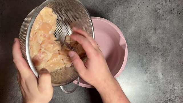 Horizontal video close up. 60 fps. Top view. Male hand putting chopped raw chicken meat pieces from metal colander to pink bowl on kitchen. Chef preparing lunch, dinner. Concept of no vegan food cafe