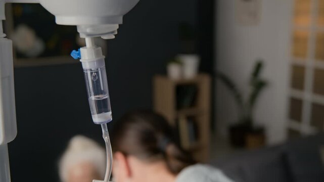 Close-up selective focus view of young female physiotherapist setting intravenous infusion system for senior woman while visiting her at home