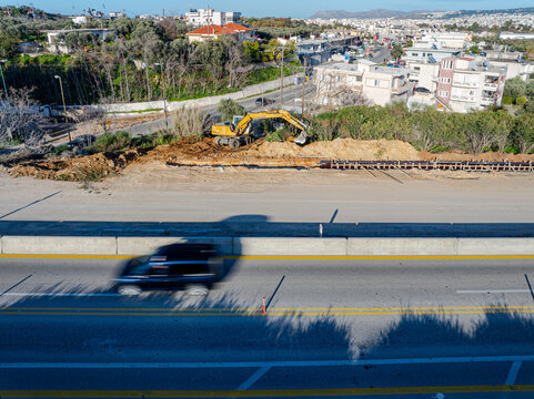Aerial view of an excavator amidst earthworks next to a busy road, with the blur of passing cars creating a dynamic contrast against the static scene, Chania, Chania, Greece.