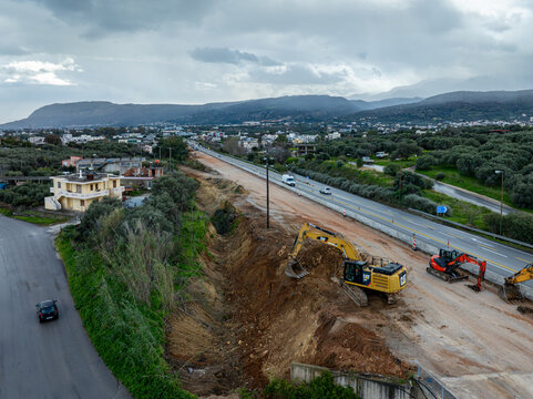Aerial view of heavy machinery reshaping the earth near a bustling highway under a cloudy sky, blending industrial activity with natural landscape, Chania, Chania, Greece.