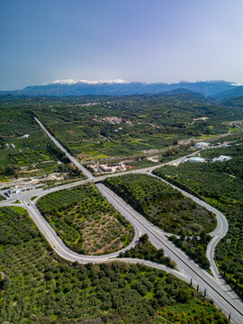 Aerial view of the serpentine highway interchange winding through the lush green landscape, framed by distant mountains under a vast blue sky, Chania, Chania, Greece.