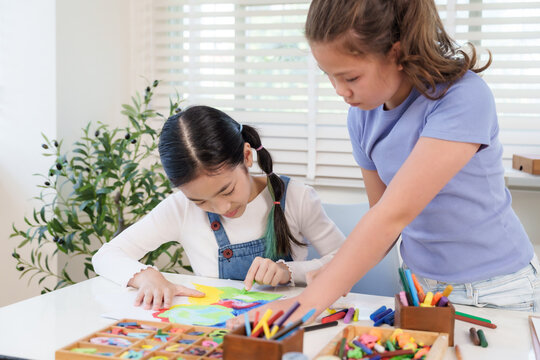 Caucasian preteen girl leaning over desk helping asian friend drawing colorful artwork during classroom elementary art education teamwork activity creativity childhood learning practice
