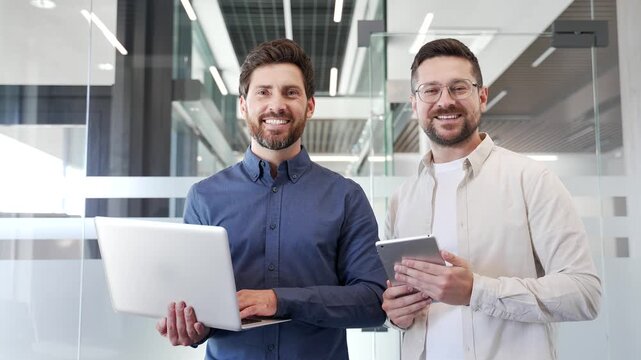 Portrait of two smiling businessmen holding laptop and digital tablet standing in modern business office. Team of happy confident successful entrepreneurs looking at camera. Teamwork of professionals