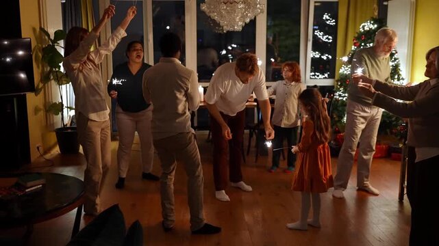 Father lights a sparkler for his daughter and a female friend gives a sparkler to a male guest while a family dances around them