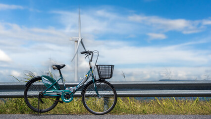 Bicycle parked beside the road with wind turbine in background under blue sky and white clouds.