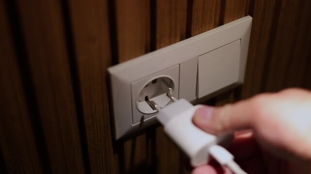 Horizontal defocused video close up. Male hand. Man inserts charger plug of mobile phone, gadget, laptop, extension cord, computer, TV in white plastic wall socket on wood wall in dark hotel room