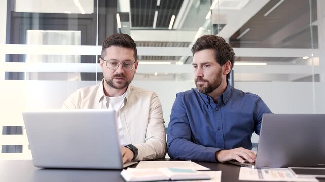 A mature employee gives advice to a colleague who works on a laptop computer while sitting at a desk at a workplace in a modern business office. Manager advises worker, consults, explains work issue
