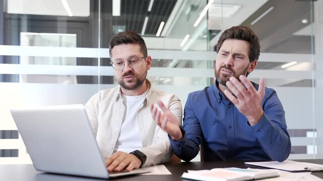 Two disappointed businessmen read bad news on laptop computer while sitting at workplace in business office. Frustrated worried colleagues co-workers are upset reviewing negative financial results