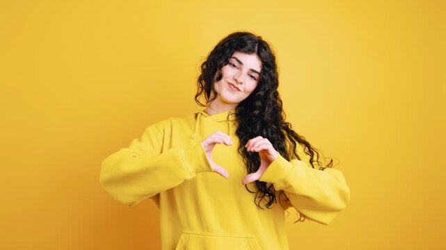 Cheerful young woman with long curly hair wearing a hoodie and showing a heart gesture with her hands, expressing love and affection