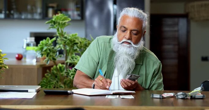 Indian mid age man counting Indian money notes at home, stylish bearded male managing monthly finance using calculator and diary while handling five hundred rupee currency notes at table