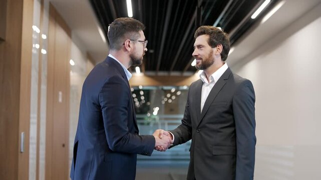 Two confident businessmen in formal suits greet each other with a handshake in a corridor of modern business center. Colleagues engaged in teamwork, communicate, discuss work issues, exchanging ideas