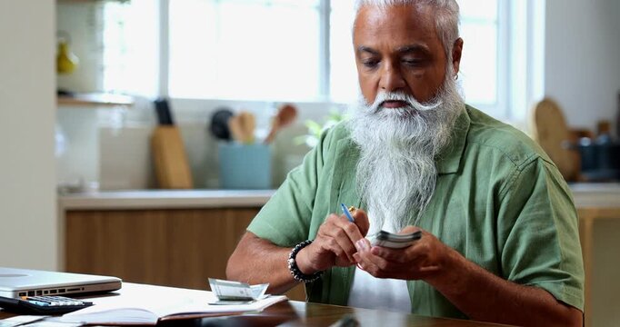 Indian mid age man counting Indian money notes at home, stylish bearded male managing monthly finance using calculator and diary while handling five hundred rupee currency notes at table