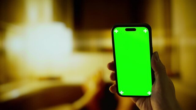 Hand holding smartphone with bright green screen mockup in warm blurred indoor environment, showcasing chroma key display for app design or video editing