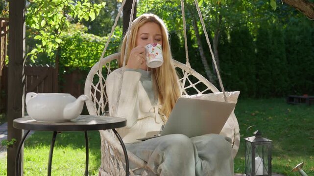 Middle aged woman working remotely outdoors on terrace with laptop and coffee mug. Cozy lifestyle, natural light, peaceful atmosphere, balance between work and life.
