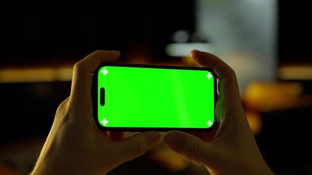 Hand holding modern smartphone with bright green screen in dimly lit interior, showcasing touchscreen display for mobile app design, video mockup, and digital media editing