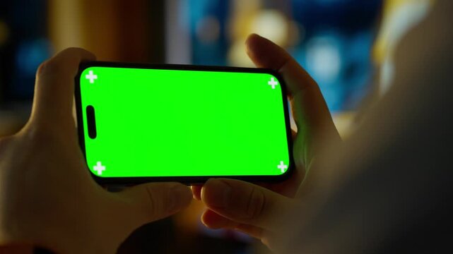 Person holding smartphone with bright green chroma key screen in cozy indoor setting, showcasing mobile mockup for app design, video editing, or user interface preview
