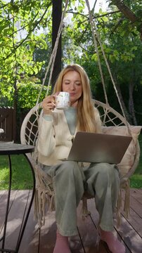 Middle aged woman working remotely outdoors on terrace with laptop and coffee mug. Cozy lifestyle, natural light, peaceful atmosphere, balance between work and life.