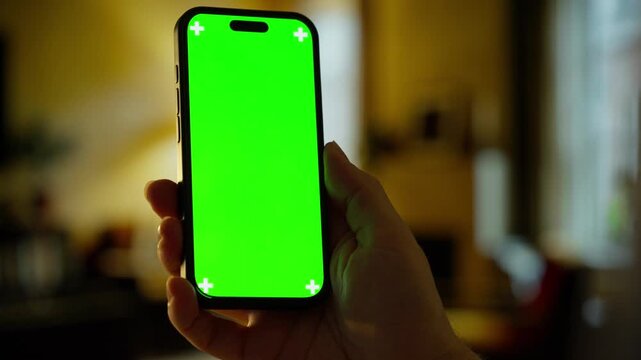 Person holding modern smartphone with bright green chroma key screen in warm indoor environment for digital compositing and mobile app presentation