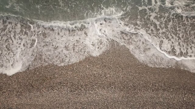 Loop-ready 4K UHD static overhead shot of foamy waves washing over a pebble beach shoreline; soothing natural texture for commercials, intros, wellness backgrounds.