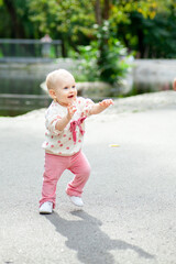 A baby is taking first steps in a park. The child is smiling and looking happy while walking on a path. Trees and a pond are visible in the background.