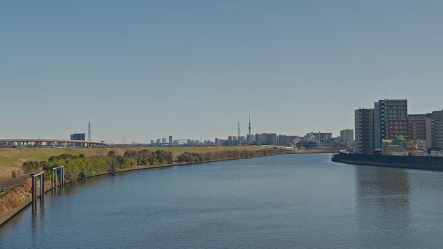 A peaceful wide-angle shot of the Sumida River in Tokyo on a clear day. The Tokyo Skytree and city skyline are visible in the far distance beyond the riverbanks and residential buildings.