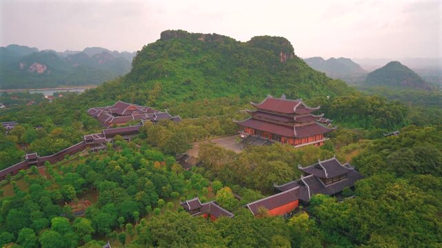 A majestic high-angle aerial view showcasing the grand Tam The Hall with its traditional multi-tiered dark tiled roofs, nestled within the lush green forests and limestone hills of the Bai Dinh Pagoda