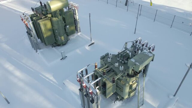Low altitude view over high voltage power distribution equipment operating in freezing snowy conditions under winter sunlight. Icy frost coating the electrical substation transformers machinery
