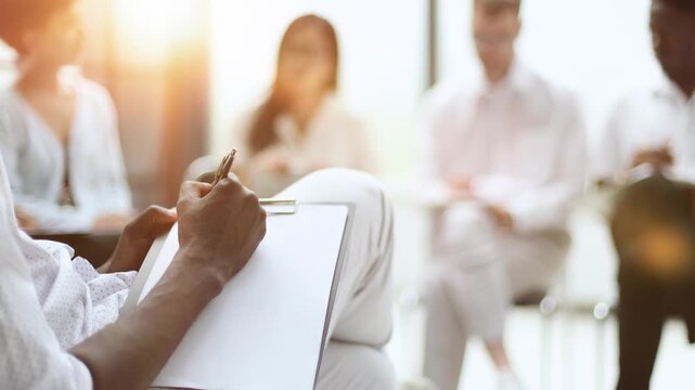 A hand writes with a pen in a notebook against the background of colleagues sitting on a chair