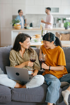Friends laughing and using laptop on couch with wine at home