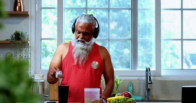 Indian mid age fit man with beard making protein shake post workout at home, stylish healthy male wearing headphones and sando vest preparing nutrition supplement in modern kitchen interior