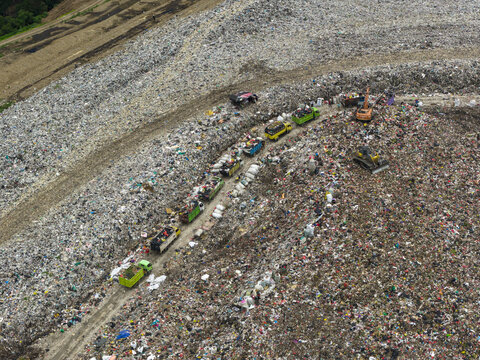 Aerial view of a vast, textured landscape of waste, where colorful trucks navigate the monochrome terrain, highlighting the scale of human impact, Denpasar, Bali, Indonesia.