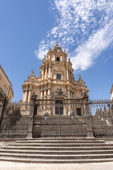Fototapeta premium Baroque cathedral facade with ornate iron gate and steps under blue sky