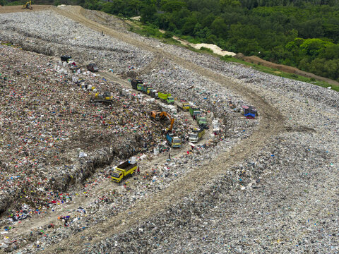 Aerial view of a vast landfill site shows a stark contrast between the vibrant green forest and the monochromatic sea of waste, with trucks navigating the mounds of trash, Denpasar, Bali, Indonesia.