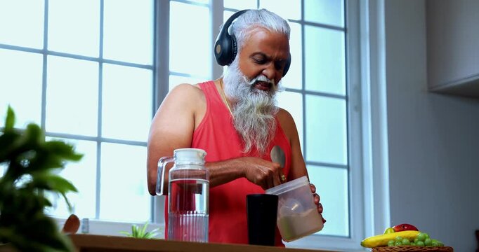 Indian mid age fit man with beard making protein shake post workout at home, stylish healthy male wearing headphones and sando vest preparing nutrition supplement in modern kitchen interior