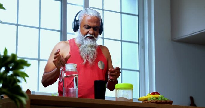 Indian mid age fit man with beard making protein shake post workout at home, stylish healthy male wearing headphones and sando vest preparing nutrition supplement in modern kitchen interior